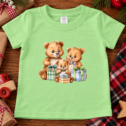 A keylime green short-sleeve baby t-shirt featuring a printed graphic of three cute teddy bears hugging and playing with wrapped Christmas presents. The shirt is displayed flat on a rustic wooden surface surrounded by holiday-themed props like a toy car, plaid fabric, and pine cones.