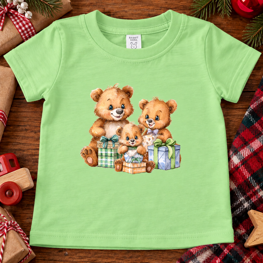 A keylime green short-sleeve baby t-shirt featuring a printed graphic of three cute teddy bears hugging and playing with wrapped Christmas presents. The shirt is displayed flat on a rustic wooden surface surrounded by holiday-themed props like a toy car, plaid fabric, and pine cones.
