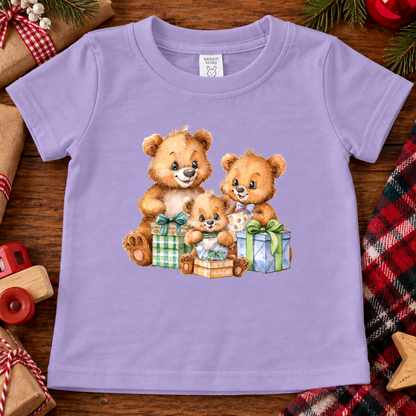 A lavender purple short-sleeve baby t-shirt featuring a watercolor-style graphic of three teddy bears hugging and opening Christmas presents. The t-shirt is displayed flat on a rustic wooden background surrounded by holiday decorations like pine branches, a red plaid scarf, a toy car, and wooden stars.