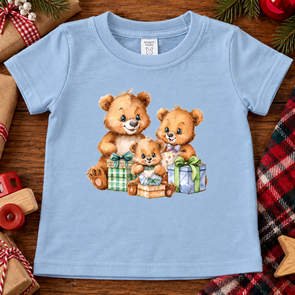 A light blue short-sleeve baby t-shirt with a watercolor graphic of three teddy bears sharing gift boxes, laid flat on a rustic wooden background with Christmas-themed props.
