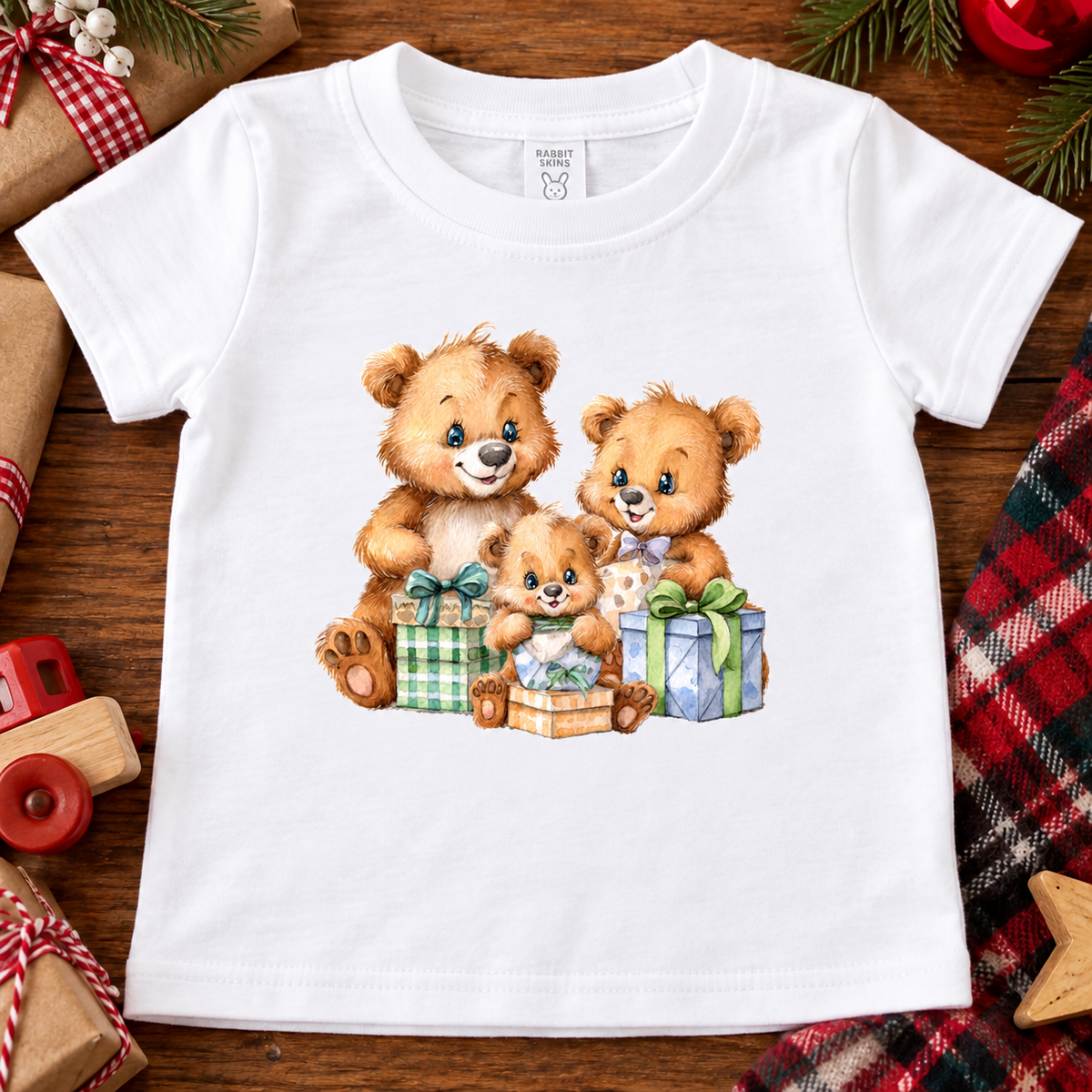 A white short-sleeve baby t-shirt with a watercolor-style graphic of three teddy bears surrounded by Christmas presents, laid flat on a wooden surface with holiday decorations.