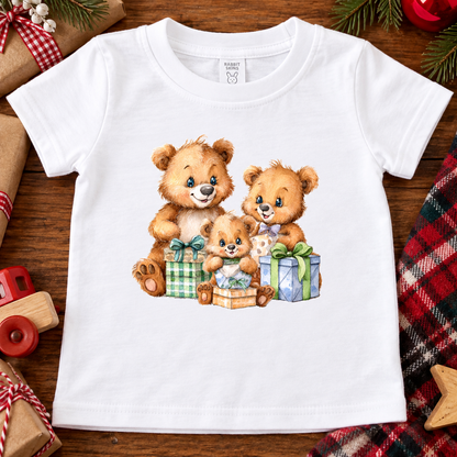 A white short-sleeve baby t-shirt with a watercolor-style graphic of three teddy bears surrounded by Christmas presents, laid flat on a wooden surface with holiday decorations.
