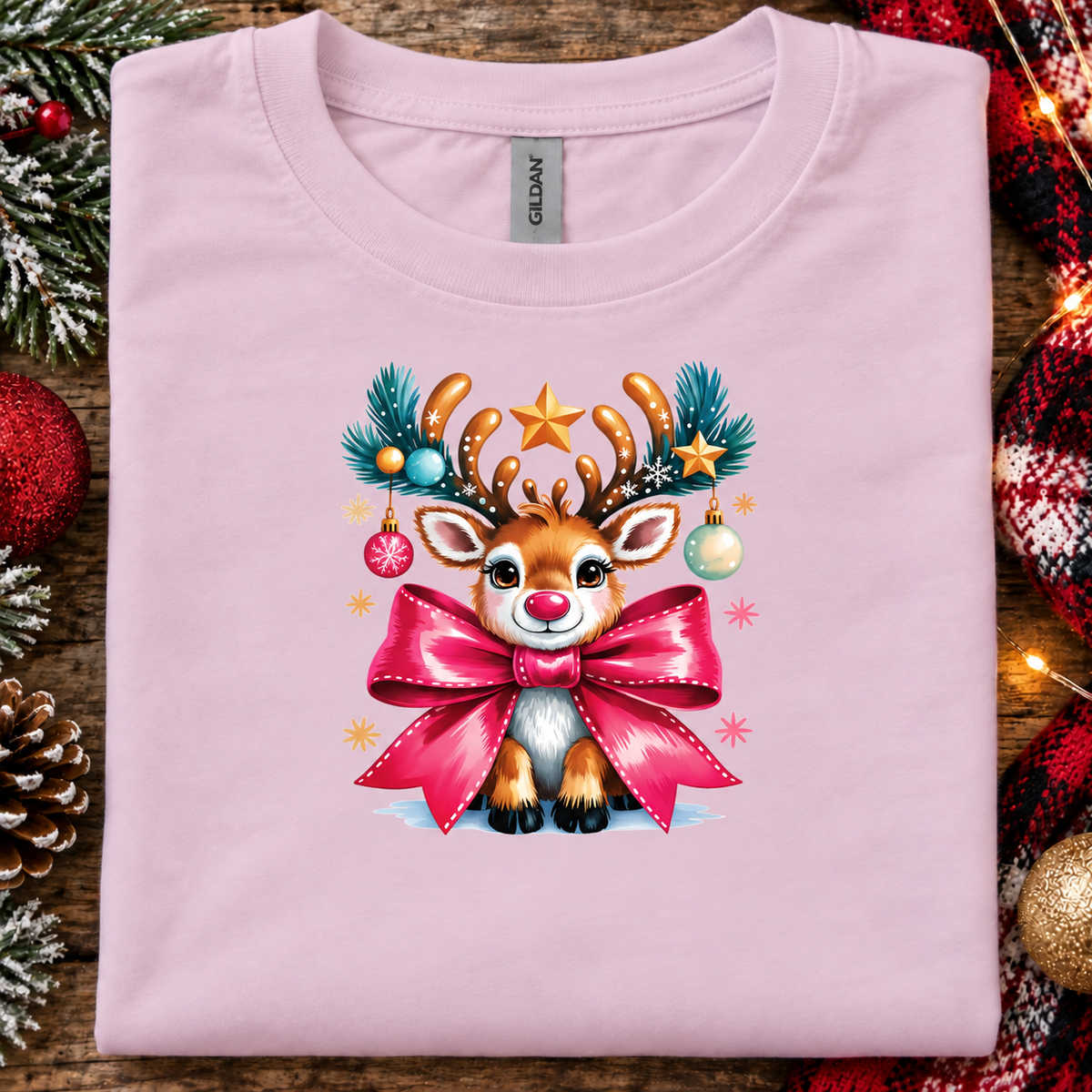 A folded light pink Gildan t-shirt featuring a colorful Christmas graphic of a reindeer with antlers decorated with pine branches, ornaments, and a large pink bow. The shirt is styled on a rustic wooden background with holiday decorations like pinecones, a red ornament, and a plaid blanket.
