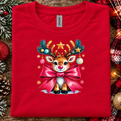 A folded bright red Gildan t-shirt with a festive Christmas graphic of a cute reindeer wearing antlers and a large pink bow, displayed on a rustic wooden background with holiday decorations.