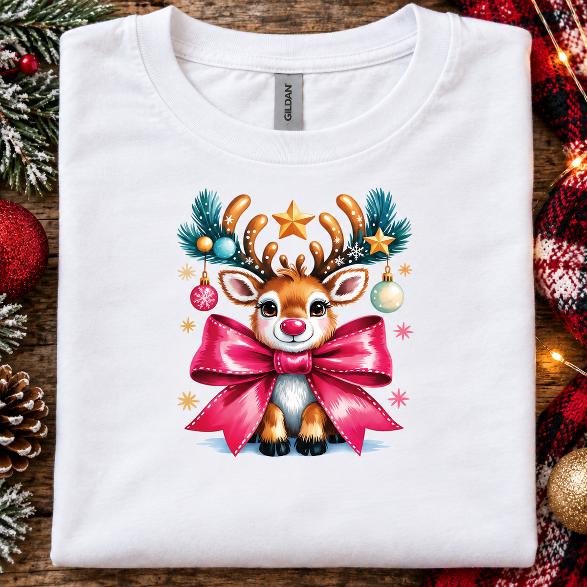 A folded white Gildan t-shirt with a colorful Christmas graphic of a cute reindeer wearing a large pink bow, displayed on a rustic wooden background with holiday decorations.