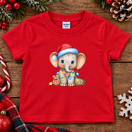 A red short-sleeve Gildan baby t-shirt featuring a cartoon Christmas elephant graphic, styled in a flat-lay on a rustic wooden background with holiday decorations.
