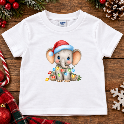 A white Gildan short-sleeve baby t-shirt featuring a cute cartoon baby elephant wearing a red Santa hat and wrapped in colorful Christmas lights, displayed on a rustic wooden background with holiday decorations.