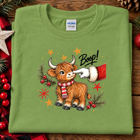 A folded kiwi green Gildan Softee t-shirt with a festive graphic of a Highland cow wearing a red plaid scarf, being gently propped by a Santa hat sleeve. The graphic includes the word 'Boop!' and holiday accents like stars, berries, and pine branches, displayed on a rustic wooden background with seasonal decorations.