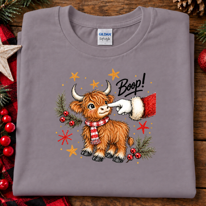 A folded paragon colored Gildan Softstyle t-shirt with a Christmas Highland cow graphic, displayed on a rustic wooden background with holiday decorations.