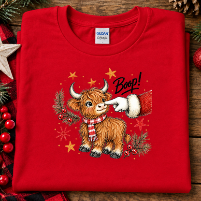 A folded red Gildan Straight crewneck sweatshirt with a festive graphic of a Highland cow wearing a red and white plaid scarf, surrounded by gold stars, pine branches, and the word 'Boop!' in black script, displayed on a rustic wooden background with holiday decorations.