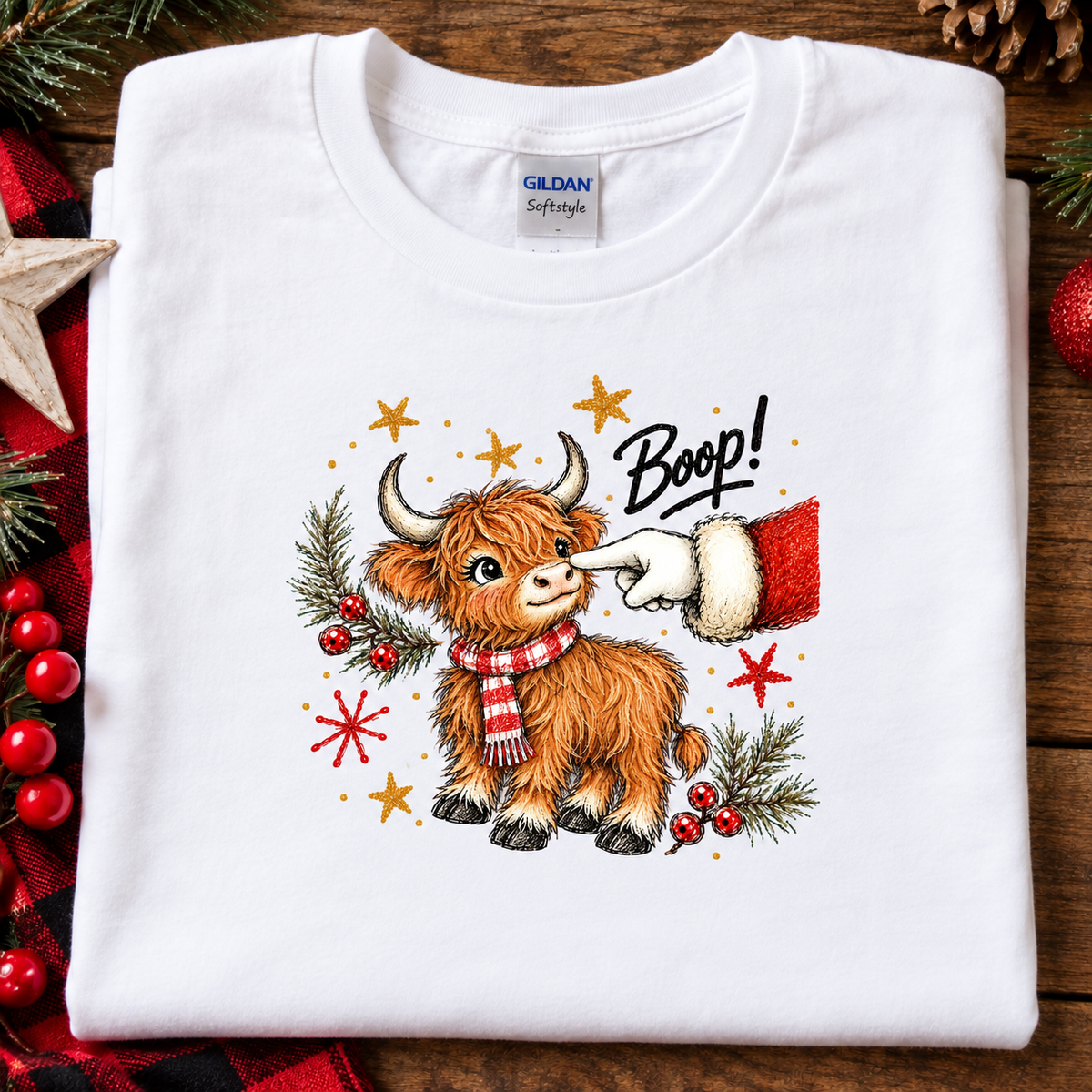 A folded white Gildan Softstyle t-shirt with a festive graphic of a Highland cow wearing a red and white striped scarf, with a Santa hat pointing at it and the word 'Boop!' written above. The shirt is displayed on a rustic wooden background decorated with Christmas elements.