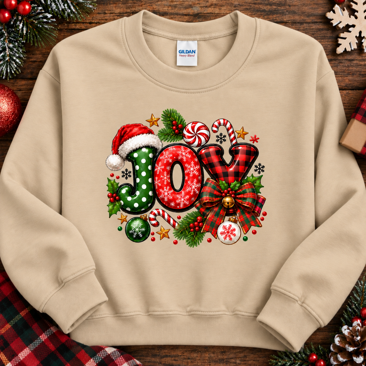 A sand colored crewneck sweatshirt with a colorful 'JOX' Christmas graphic, laid flat on a dark wooden background decorated with holiday elements like pine branches, ornaments, and a gift box. The brand label 'GILDAN' is visible inside the collar.