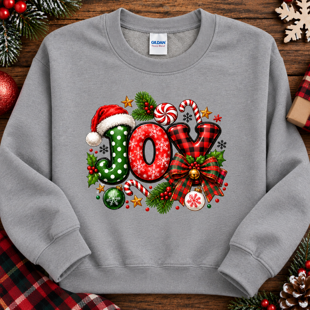 A light gray crewneck sweatshirt featuring a festive Christmas 'JOY' graphic with candy canes, a Santa hat, plaid, and ornaments, laid flat on a wooden background with holiday decorations.
