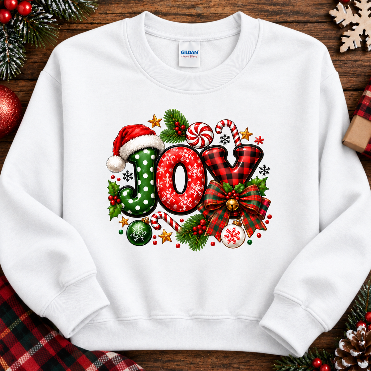 A white crewneck sweatshirt featuring a colorful 'JOY' graphic with Christmas motifs like candy canes, ornaments, pinecones, and a Santa hat, laid flat on a rustic wooden background with holiday decorations.