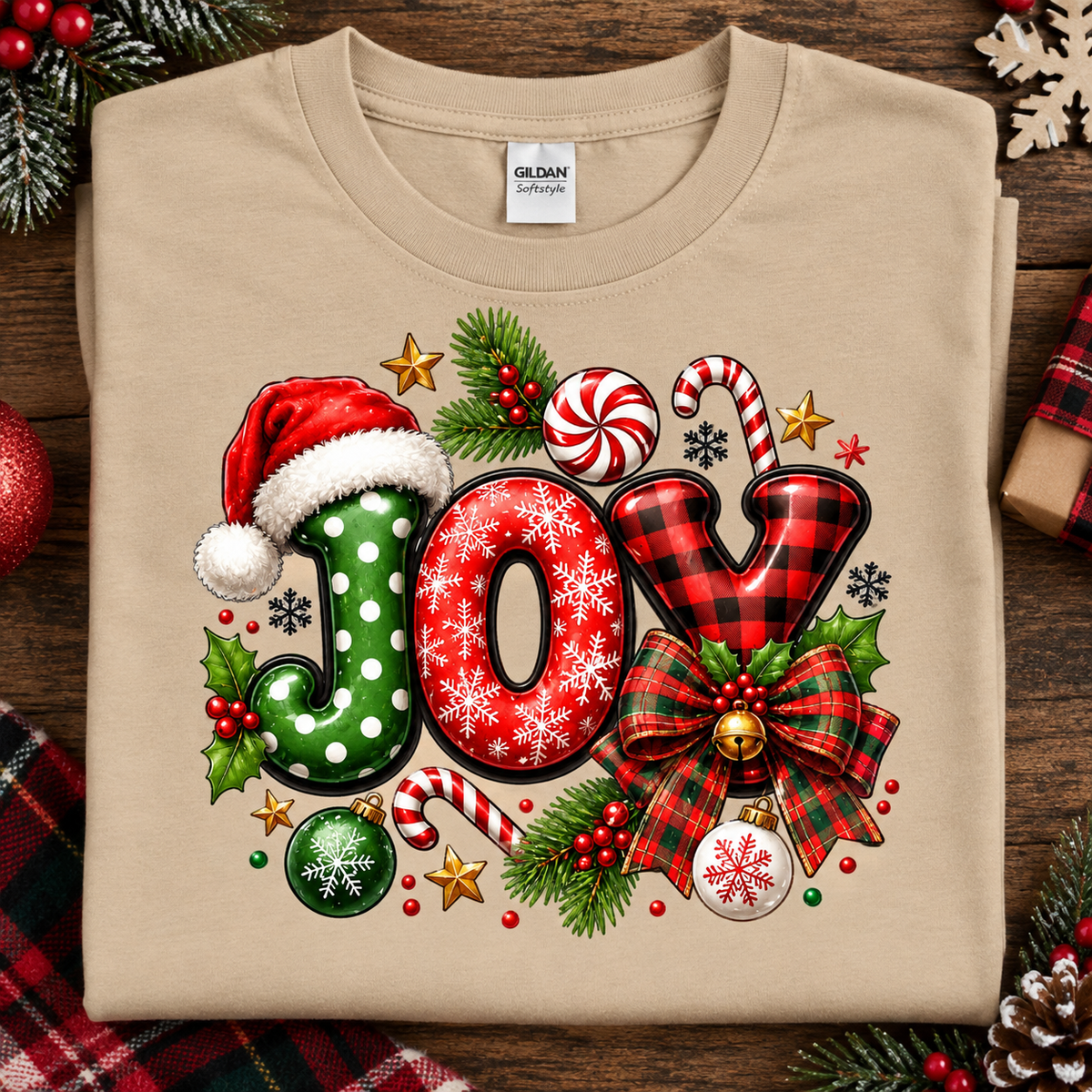 A sand colored Gildan Comfort T-shirt laid flat on a rustic wooden surface, decorated with a large festive 'JOY' graphic in red, green, black, white, and holiday colors. The shirt is surrounded by seasonal props including pine branches, ornaments, candy canes, and plaid fabric.
