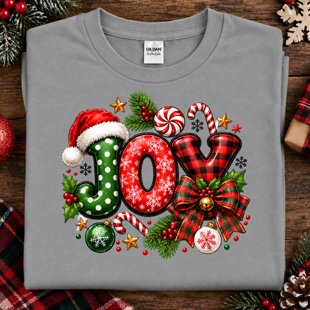 A folded light gray Gildan Comfort T-shirt laid flat on a rustic wooden surface, featuring a large festive Christmas 'JOY' graphic with red, green, black, white, gold, and red plaid holiday motifs including candy canes, holly, ornaments, and a Santa hat.