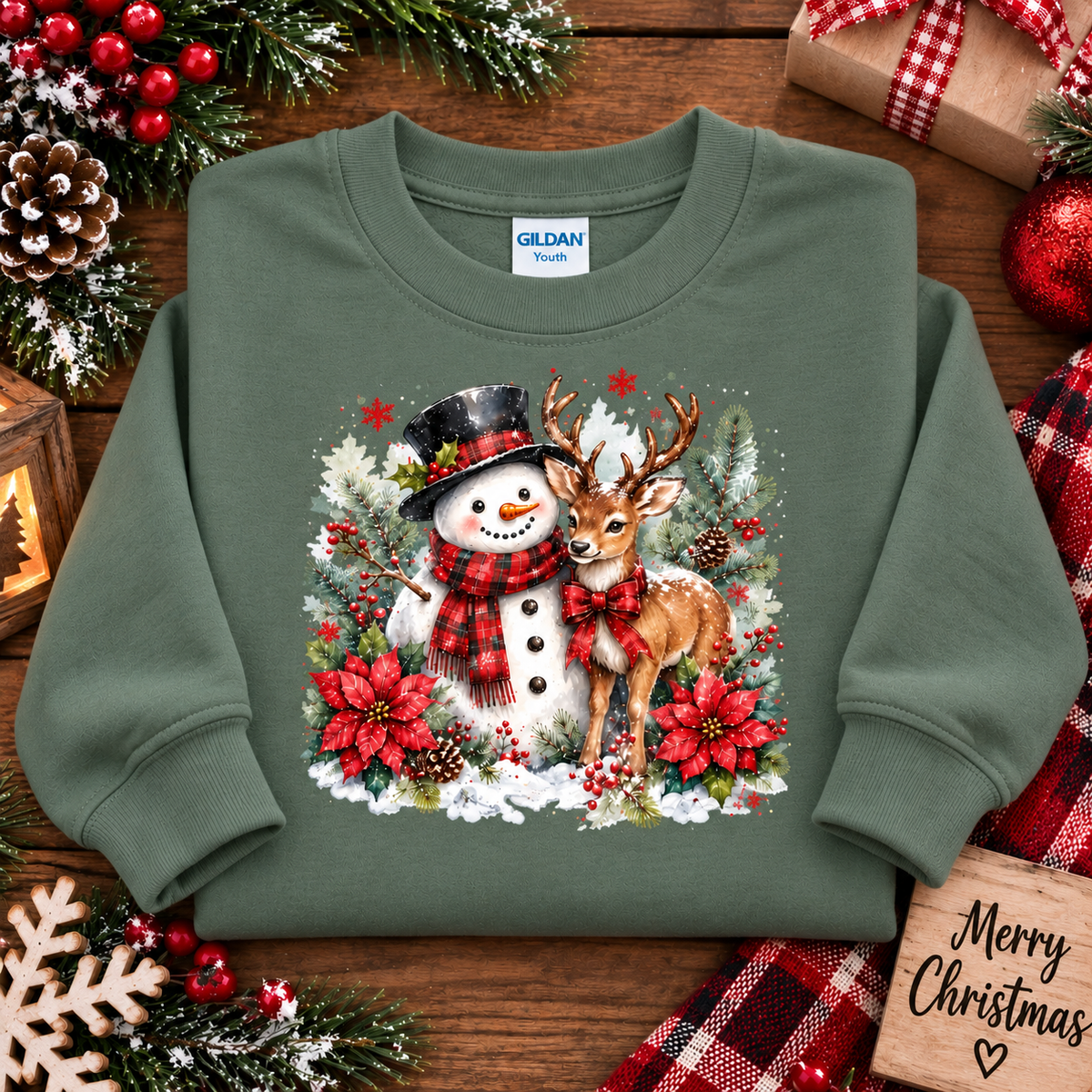 An military green Gildan Youth crewneck sweatshirt featuring a festive Christmas graphic of a snowman and a deer surrounded by poinsettias and pinecones, displayed on a rustic wooden background with holiday decorations.