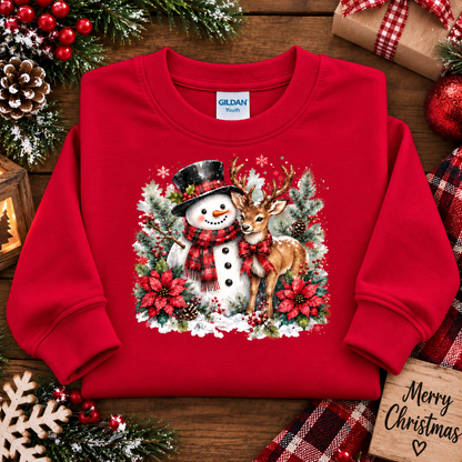 A red long-sleeve crewneck sweatshirt laid flat on a rustic wooden surface, featuring a large Christmas graphic of a snowman and a deer surrounded by pine branches and red poinsettias. The sweatshirt is styled with holiday props including a wrapped gift, pinecones, evergreen branches, and a wooden sign that reads 'Merry Christmas'.