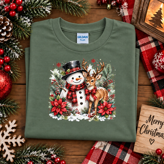 An military green Gildan Youth t-shirt is folded and presented on a rustic wooden table surrounded by Christmas decorations, including pine branches, red berries, a plaid blanket, and a wooden 'Merry Christmas' sign. The t-shirt features a colorful graphic of a snowman and a deer surrounded by poinsettias.