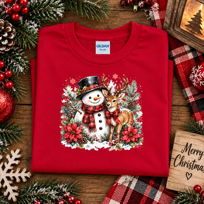A folded red Gildan Youth t-shirt with a festive Christmas snowman and deer graphic, displayed on a wooden surface surrounded by holiday decorations like pine branches, red berries, and a plaid blanket.