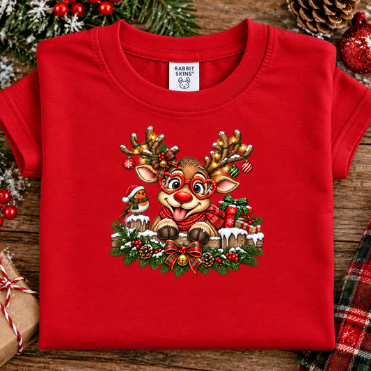 A folded red short-sleeve T-shirt with a large Christmas reindeer graphic on the front, displayed on a rustic wooden surface with holiday decorations around it. The neck label reads Rabbit Skin.