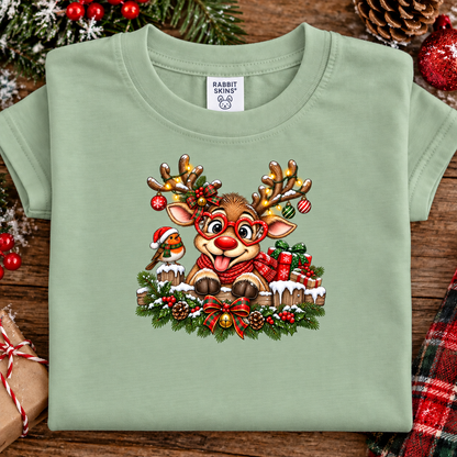 A sage green short-sleeve t-shirt with a colorful Christmas graphic of a reindeer, bird, and holiday wreath, laid flat on a rustic wooden surface with seasonal decorations.