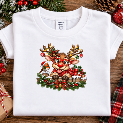 A folded white short-sleeve T-shirt with a colorful Christmas reindeer graphic on the front, displayed on a rustic wooden surface with holiday decorations including pine branches, ornaments, and a plaid fabric.