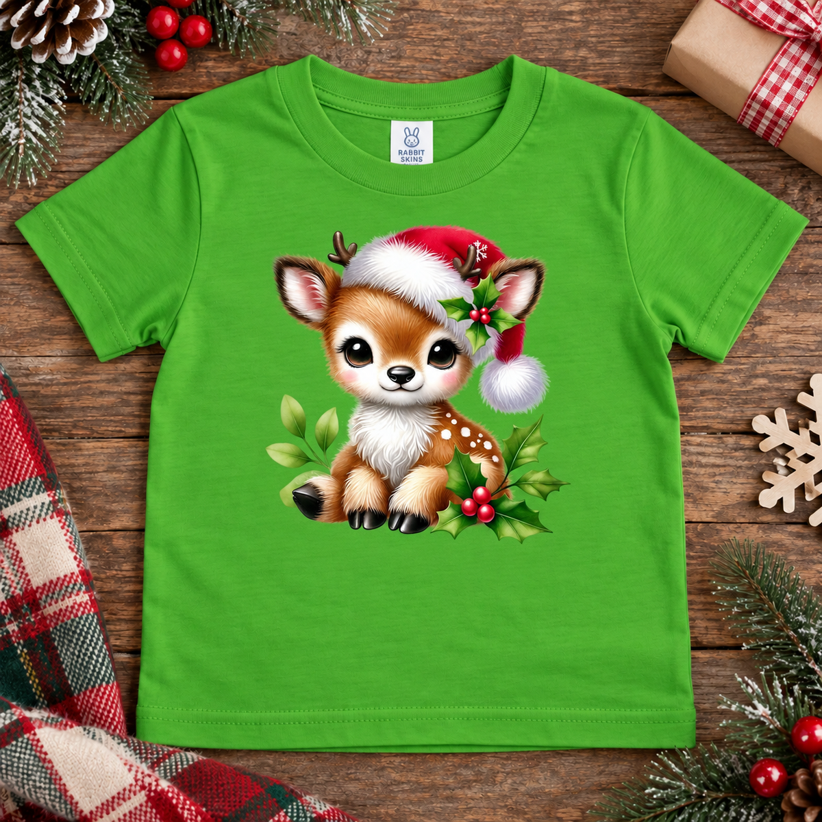 A bright green short-sleeve kids' t-shirt featuring a cute cartoon deer wearing a Santa hat and surrounded by holly, laid flat on a rustic wooden surface with holiday decorations.