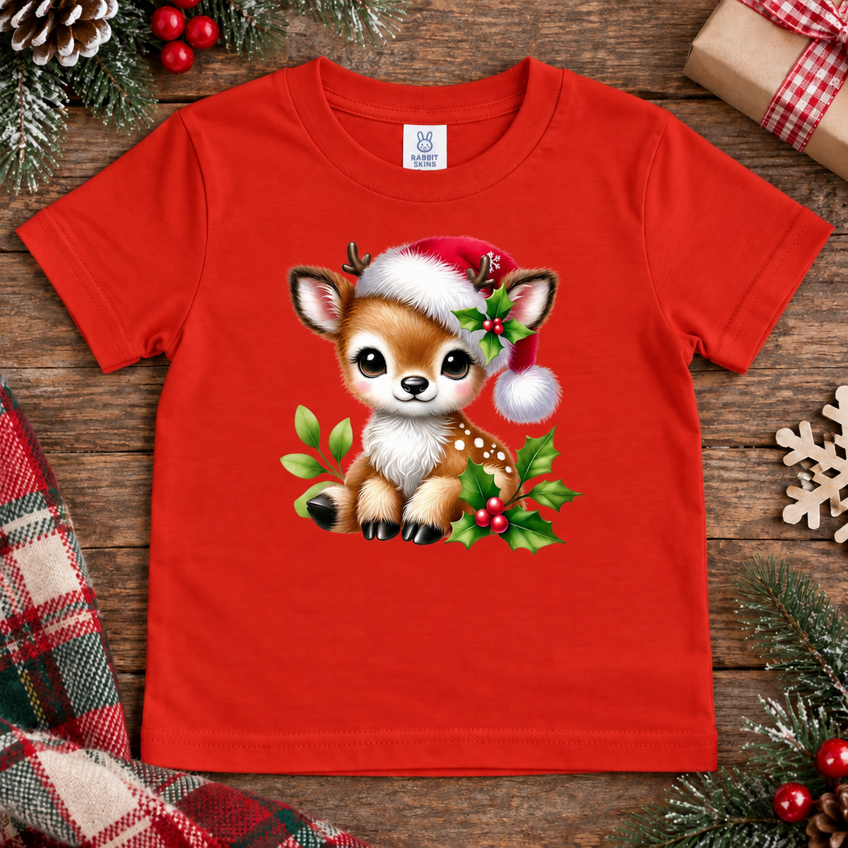 A bright red short-sleeve kids' t-shirt with a cute cartoon Christmas fawn graphic wearing a Santa hat, laid flat on a rustic wooden surface surrounded by holiday decorations.