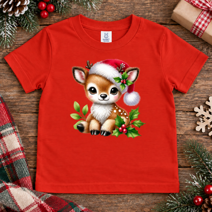 A bright red short-sleeve kids' t-shirt with a cute cartoon Christmas fawn graphic wearing a Santa hat, laid flat on a rustic wooden surface surrounded by holiday decorations.
