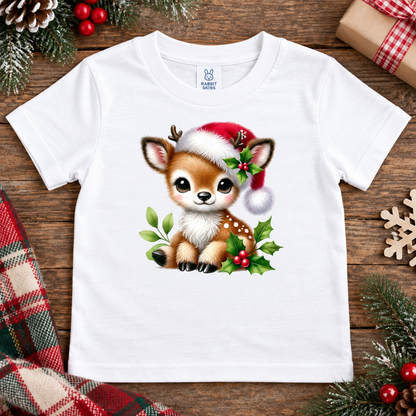 A white short-sleeve baby t-shirt with a cute cartoon Christmas fawn graphic wearing a Santa hat, displayed in a flat-lay holiday setting on a wooden surface.