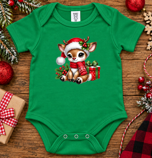 A green baby bodysuit with a festive Christmas graphic of a reindeer wearing a Santa hat and scarf, sitting next to a gift box. The bodysuit is displayed in a flat-lay arrangement on a wooden surface surrounded by holiday decorations like ornaments, pine branches, and wrapped gifts.