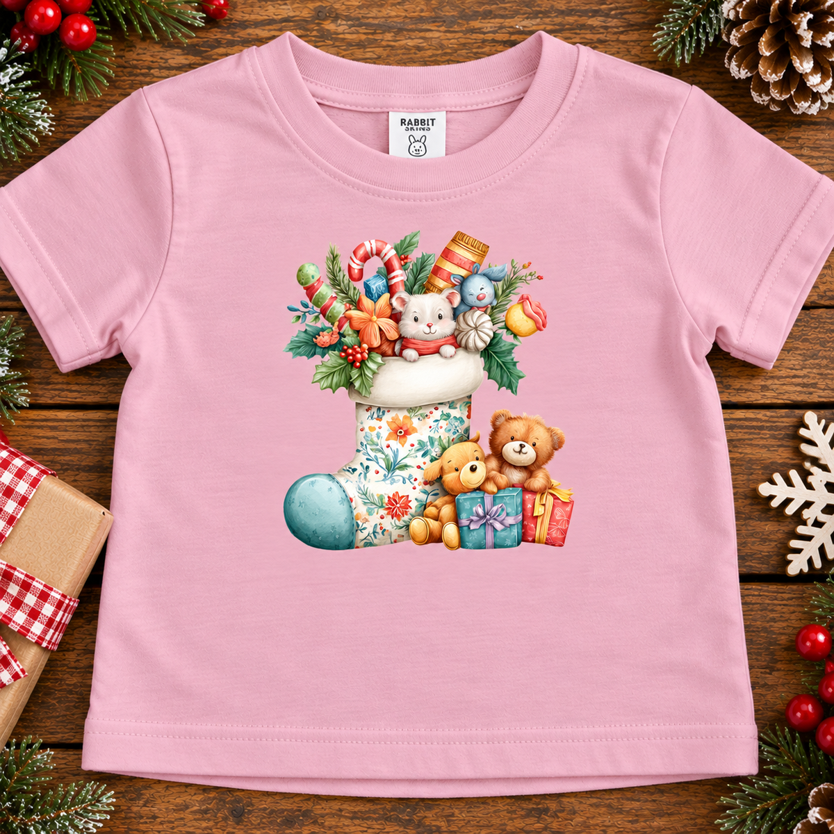 A pink short-sleeve baby t-shirt with a colorful Christmas-themed graphic print featuring a stocking, teddy bears, gifts, and candy canes, laid flat on a rustic wooden background with holiday decorations.