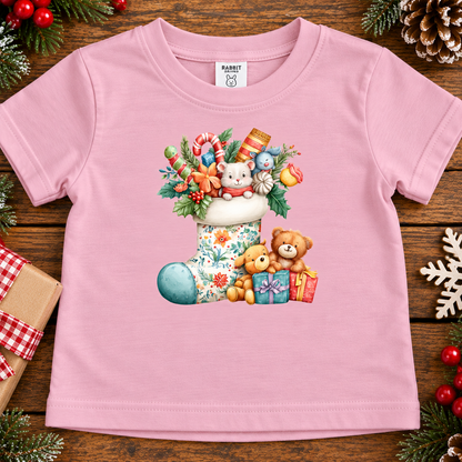 A pink short-sleeve baby t-shirt with a colorful Christmas-themed graphic print featuring a stocking, teddy bears, gifts, and candy canes, laid flat on a rustic wooden background with holiday decorations.