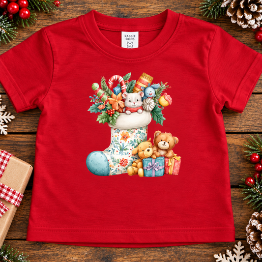 A red short-sleeve baby t-shirt with a colorful Christmas teddy bear graphic, laid flat on a rustic wooden background decorated with holiday greenery and ornaments.