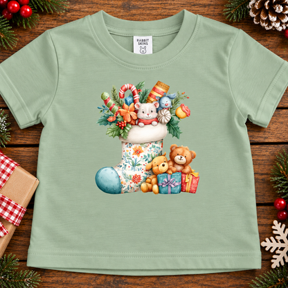 A light sage green baby t-shirt with a colorful Christmas graphic of a floral stocking, teddy bears, candy canes, and ornaments, displayed on a rustic wooden background with holiday decorations.