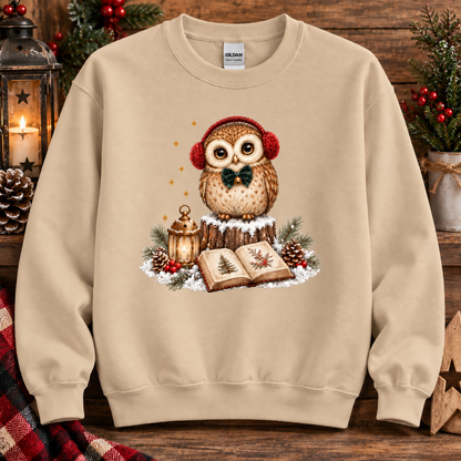A sand colored crewneck sweatshirt featuring a festive Christmas owl graphic on the chest. The graphic shows a brown owl wearing red earmuffs and a black bow tie, sitting on a tree stump next to an open book, a lantern, and pinecones.