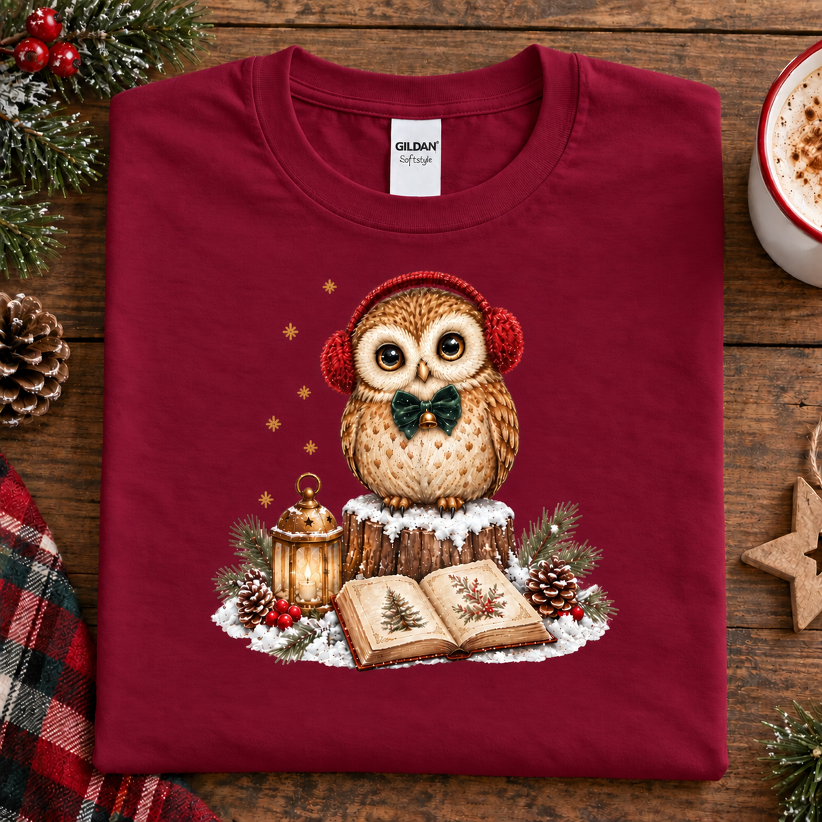 A folded cardinal red Gildan t-shirt with a Christmas owl graphic, displayed on a rustic wooden table surrounded by holiday decorations like pine branches, a pinecone, a star ornament, and a mug of hot cocoa.