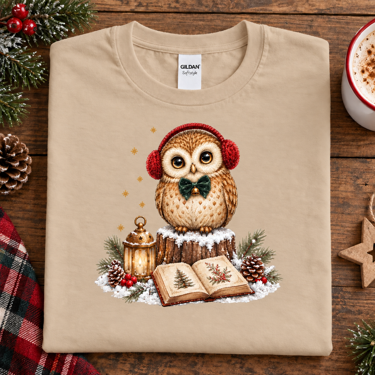 A sand colored Gildan t-shirt with a Christmas owl graphic, laid flat on a rustic wooden table surrounded by holiday decorations like pine branches, a mug of coffee, and a plaid cloth.