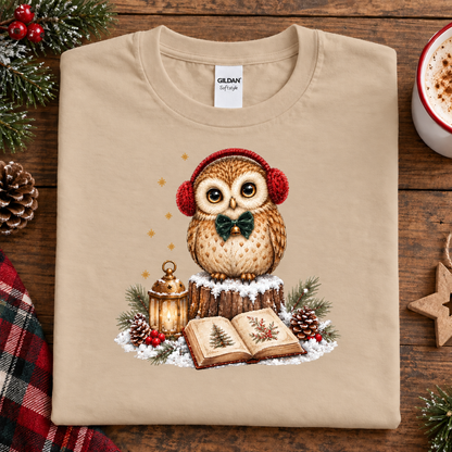 A sand colored Gildan t-shirt with a Christmas owl graphic, laid flat on a rustic wooden table surrounded by holiday decorations like pine branches, a mug of coffee, and a plaid cloth.