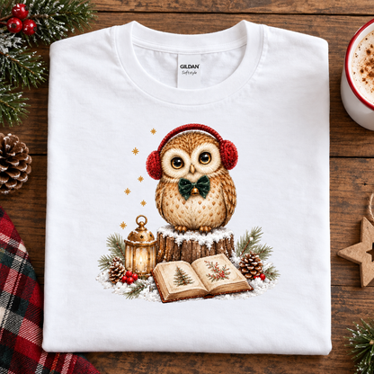 A white Gildan t-shirt laid flat on a rustic wooden table, featuring a festive Christmas owl graphic wearing red earmuffs and a green bow tie, surrounded by a lantern, open book, pinecones, and evergreen branches.