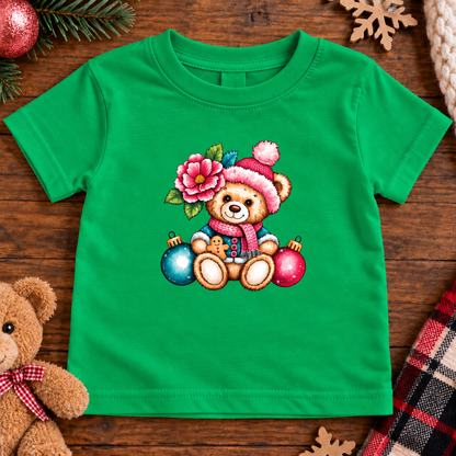 A bright green short-sleeve baby t-shirt with a festive Christmas teddy bear graphic, styled in a flat-lay on a dark wooden background with holiday props.