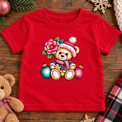 A red short-sleeve baby t-shirt with a colorful graphic of a teddy bear wearing a Santa hat and scarf, surrounded by a pink flower and Christmas ornaments, laid flat on a wooden surface with holiday decorations.