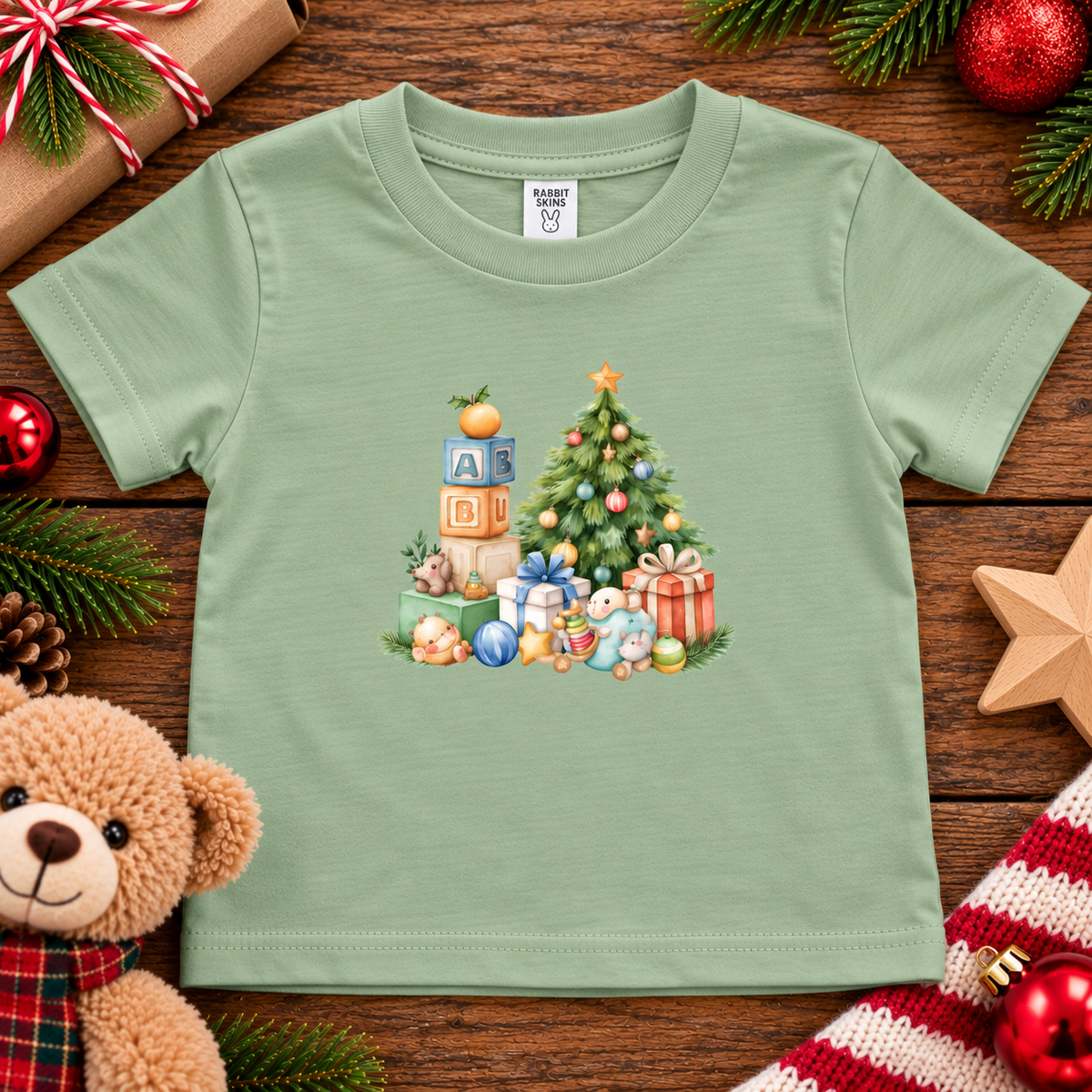 A sage colored toddler t-shirt with a Christmas graphic with a tree, blocks and presents