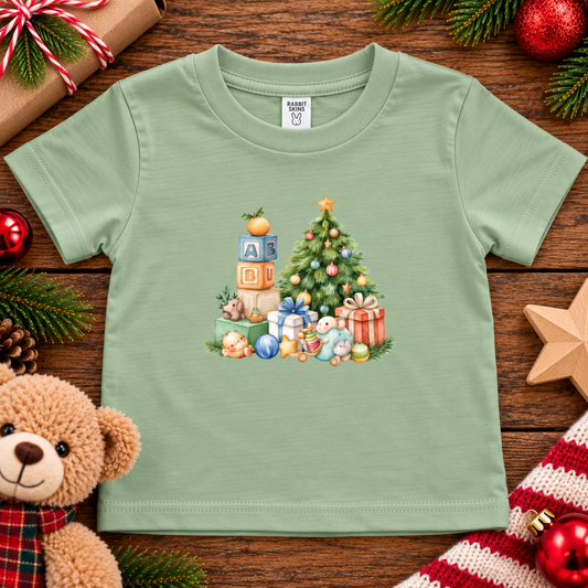 A sage colored toddler t-shirt with a Christmas graphic with a tree, blocks and presents