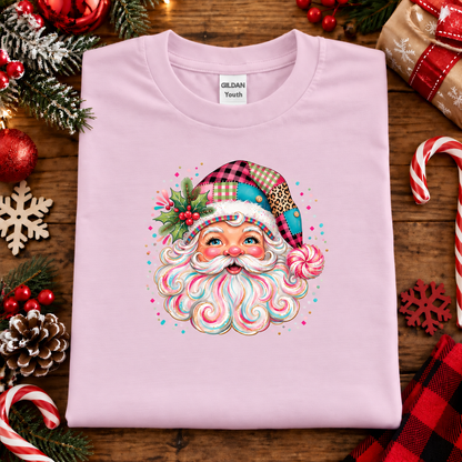 A folded light pink Gildan Youth t-shirt with a colorful Christmas Santa graphic, displayed on a rustic wooden background decorated with holiday ornaments, candy canes, pinecones, and a wrapped gift.