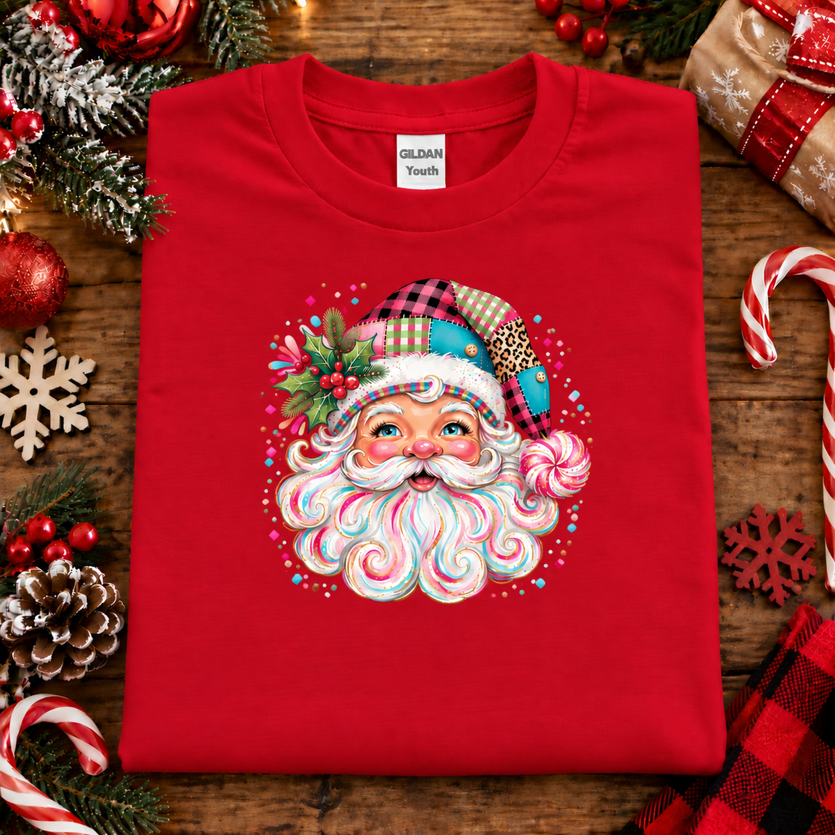 A folded red Gildan Youth t-shirt featuring a colorful Christmas-themed Santa graphic, styled on a rustic wooden background with holiday decorations like candy canes, pinecones, and ornaments.