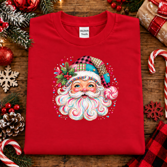 A folded red Gildan Youth t-shirt featuring a colorful Christmas-themed Santa graphic, styled on a rustic wooden background with holiday decorations like candy canes, pinecones, and ornaments.