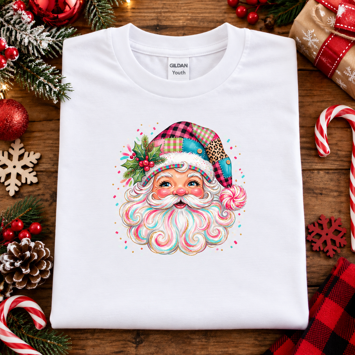 A folded white Gildan Youth t-shirt with a colorful Christmas Santa graphic, displayed on a rustic wooden background surrounded by holiday decorations like candy canes, pinecones, and ornaments.
