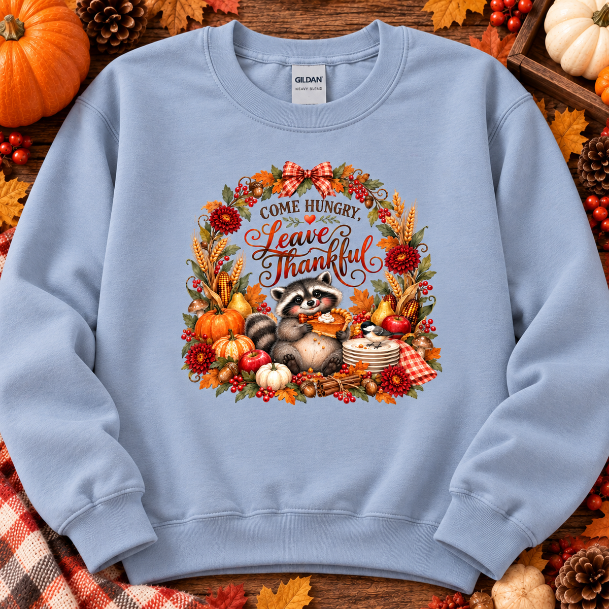 A light blue crewneck sweatshirt featuring a colorful autumn-themed graphic with a raccoon, pumpkins, leaves, and the text 'COME HUNGRY. Leave Thankful.